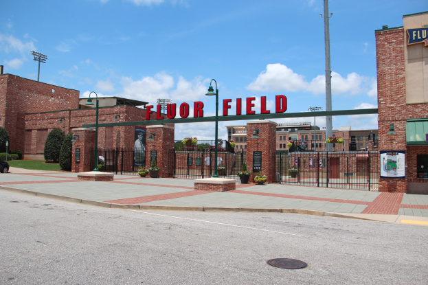 Baseballstadion-Eingangsschild mit der Aufschrift "Floor Field" umgeben von städtischen Elementen wie Gebäuden, Straßenmöbeln, Fahrzeugen, Grünflächen und einem bewölkten Himmel.