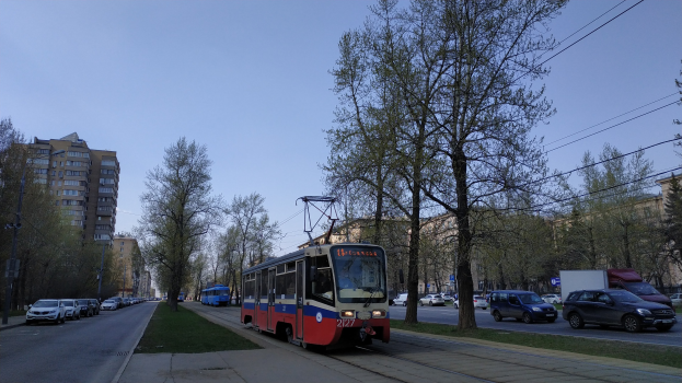 Rote und blaue Straßenbahn auf einer Stadtstraße mit parkenden Autos, Bäumen, Gebäuden und Himmel im Hintergrund, Text unten.