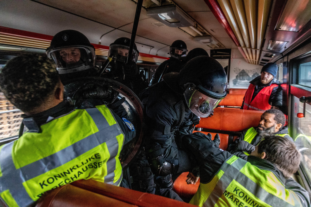 Eine Gruppe von Polizisten in Einsatzausrüstung auf einem Bus, mit einer Person in der Mitte sitzend und einem Plakat im Hintergrund.