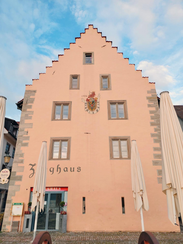 Pinkes Gebäude mit einem zentralen Uhrenturm, umgeben von Fenstern, Schirmen, einem Schild, einer Straßenlaterne, Topfpflanzen und einem bewölkten Himmel, repräsentiert Sehenswürdigkeiten in München, Deutschland.
