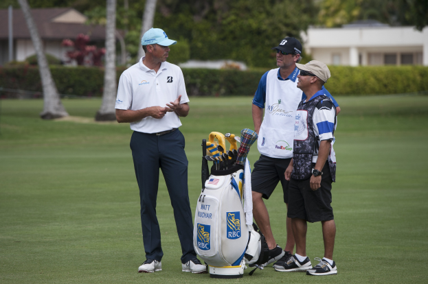 Eine Gruppe Männer steht auf einem grünen Feld mit einem Golfbag im Vordergrund, Bäume, Pflanzen und Gebäude im Hintergrund, was auf ein Golfturnier hindeutet.