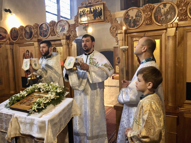 Eine Gruppe von Menschen steht um einen Tisch in einer Kirche, hält Bücher in den Händen, mit einem Tuch und einem Blumenstrauß auf dem Tisch und Fotorahmen, Lampen, einem Fenster und einem hölzernen Objekt im Hintergrund.
