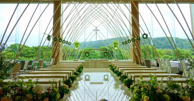 Eine Hochzeitszeremonie in einem großen Innenraum mit halbrunder Sitzordnung, Blumenschmuck und einem Blick auf Bäume, Hügel und einen klaren Himmel im Hintergrund.