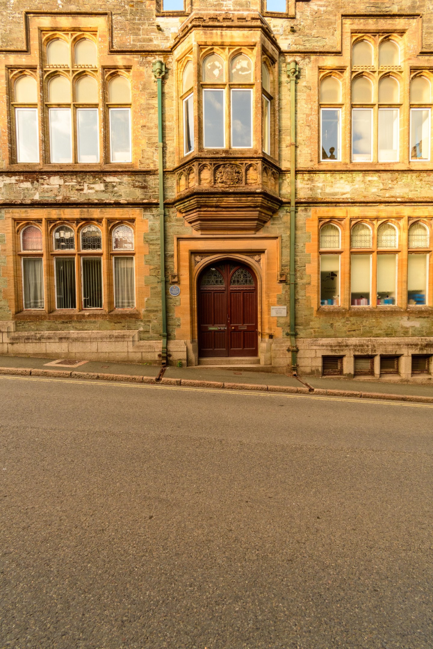 Alter Bau mit vielen Fenstern und einer hölzernen Tür, neben einer Straße gelegen, mit Rohren an seinen Wänden.