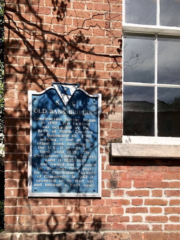 Altes Bankgebäude mit blauer Plakette an der Seite, Fenster rechts und Baum links.