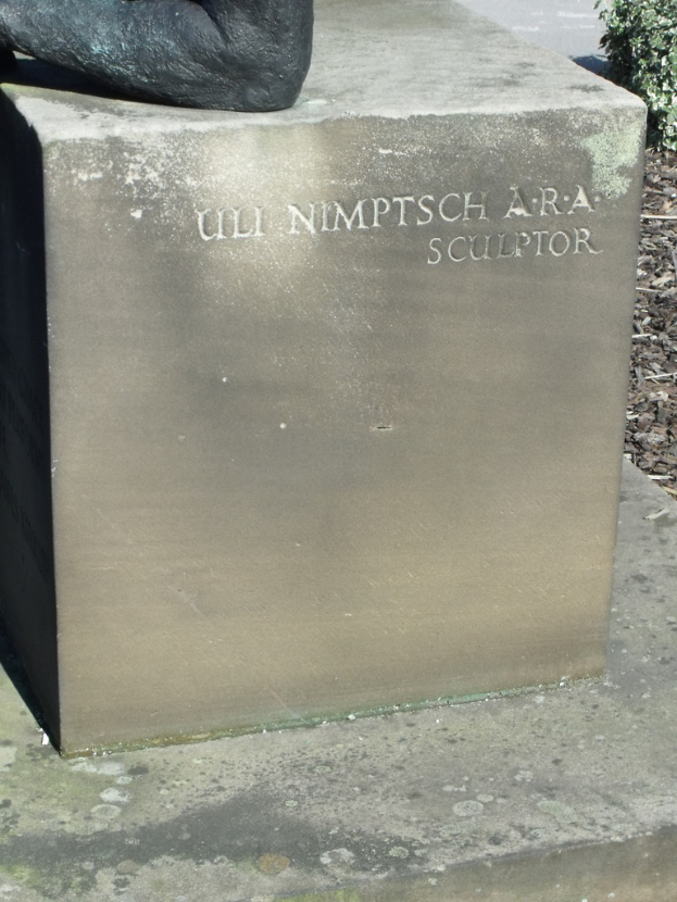 Ein Stein mit Text in Vordergrund, möglicherweise eine Statue oben, trockene Zweige, Erde und Pflanzen rechts und eine Straße im Hintergrund.