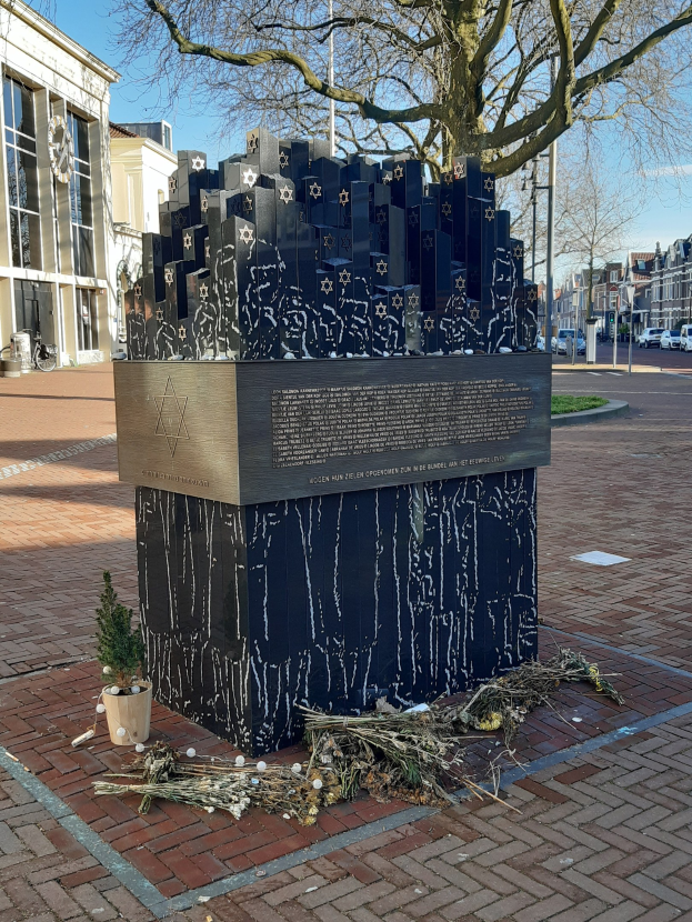Denkmal in einem Stadtplatz gewidmet an Holocaust-Opfern, mit einem Baum davor, einer Topfpflanze, verstreuten trockenen Blättern und umliegenden Gebäuden, Fahrzeugen, Pfählen und Bäumen.