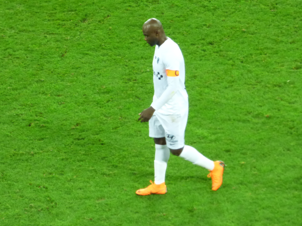 Ein Fussballspieler in einem weissen T-Shirt, weissen Shorts und orangen Schuhen überquert ein saftig grünes Feld.