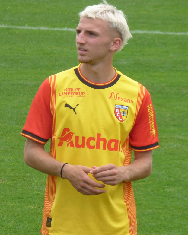 Ein Fußballspieler in einem gelb-orangen gestreiften Trikot steht selbstbewusst auf einem Feld und schaut entschlossen nach vorne.