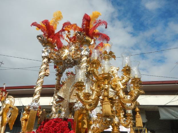 Ein großer goldener und roter Festwagen, der mit Blumen und anderen Dekorationen geschmückt ist, in einem Karnevalsumzug, mit einem Gebäude, Strommasten mit Drähten und einem bewölkten Himmel im Hintergrund.