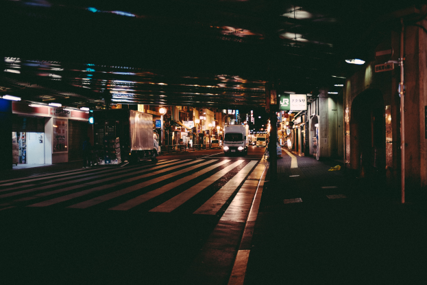 Eine nächtliche Straßenansicht mit Fahrzeugen auf der Straße, Gebäuden auf beiden Seiten, beleuchteter Gegend und einer Brücke im Hintergrund.