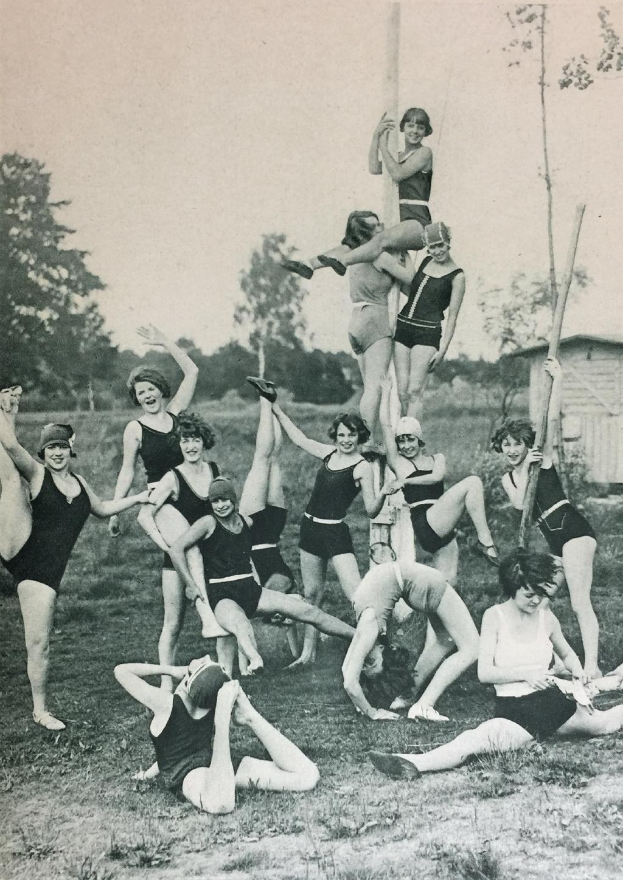 Eine Gruppe von Frauen in Badeanzügen aus den 1920er Jahren posiert für ein Foto auf einer Wiese, umgeben von Bäumen und einem Haus im Hintergrund, schwarz-weiß.