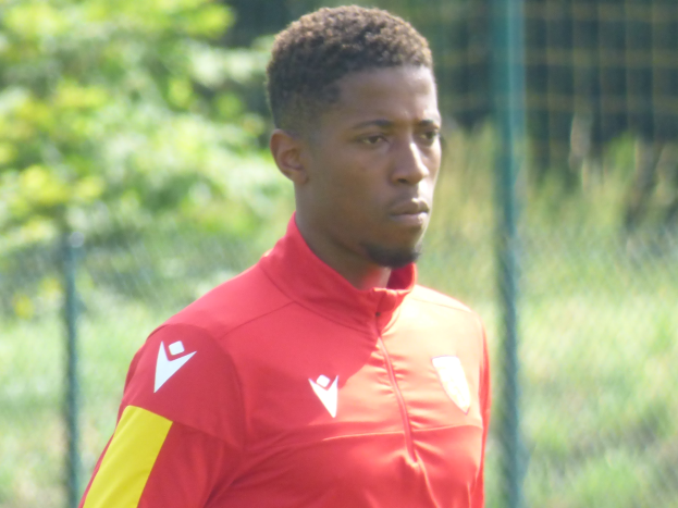 Ein junger Mann in einem roten und gelben Trikot steht auf einem Fußballfeld umgeben von einem Zaun und Bäumen und sieht entschlossen aus.