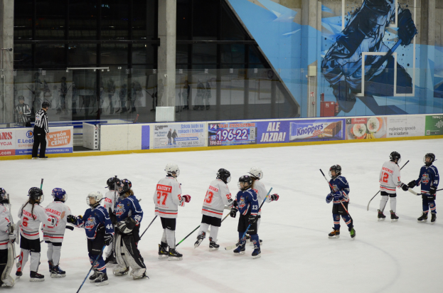 Junge Menschen in Helmen und Eishockey-Ausrüstung spielen Eishockey auf einer Indoor-Eisfläche mit Bannern, Säulen und Glaswänden im Hintergrund.