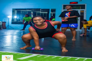 Eine Frau in einem Fitnessstudio führt eine Kniebeuge aus, während sie Hanteln hält, mit verschiedenen Fitnessgeräten und Logos im Hintergrund.