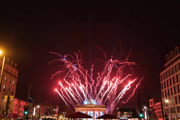 Eine belebte Stadtstraße an Silvester in Berlin, voller Menschen, Fahrzeuge und Gebäude, erleuchtet von Lichtern und Feuerwerk am Himmel.