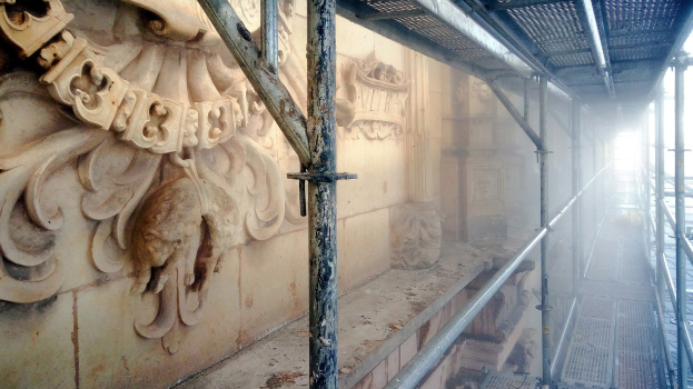 Ein renoviertes Gebäude mit detaillierten Schnitzereien an der linken Wand, Metallstangen auf der rechten Seite und einem Gerüst drumherum.