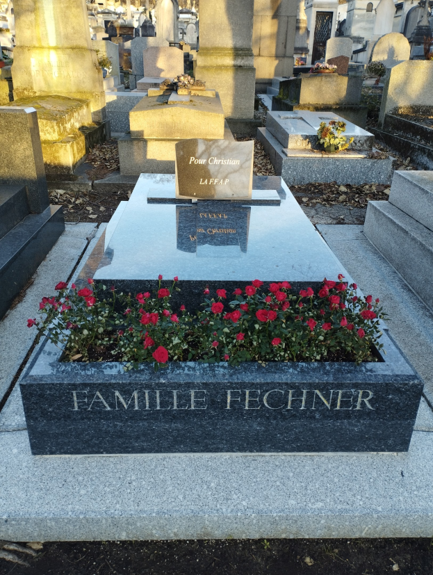 Ein Friedhof mit einem zentralen Grab, das mit roten Blumen geschmückt ist und von Bäumen umgeben ist.