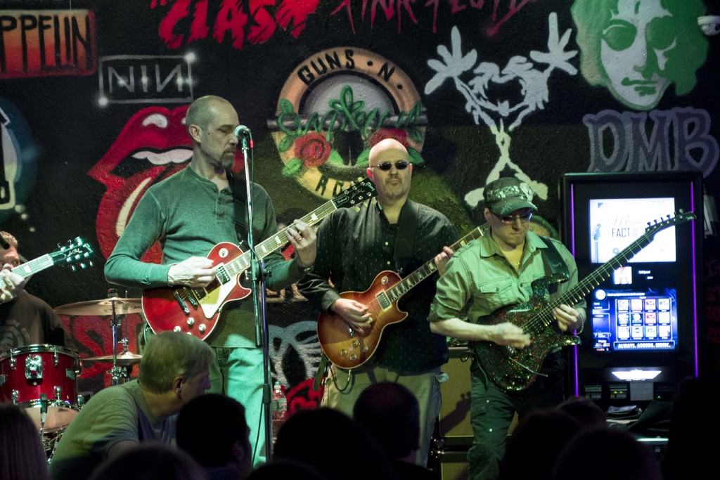 Drei Männer auf einer Bühne, die Gitarren spielen, mit einer Menge unten und einem Graffiti oder einer Wandmalerei im Hintergrund.