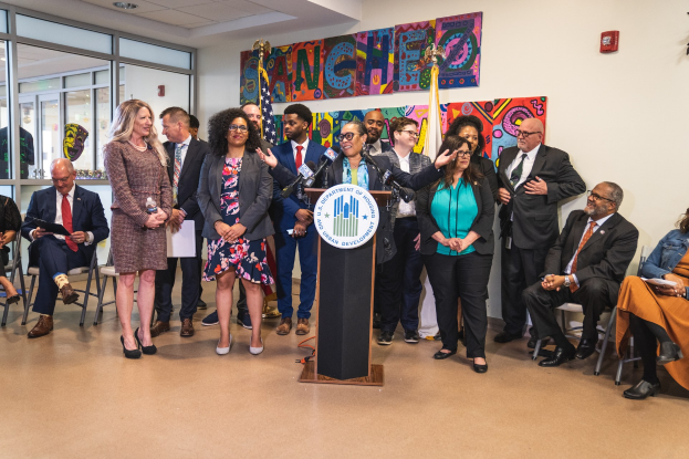 Eine Gruppe von Menschen steht vor einem Podium mit einem Mikrofon und einer Tafel mit dem Text "Sancho City Councilman Joins City Council", während andere auf Stühlen sitzen oder in der Nähe stehen; Flaggen, ein Feuerlöscher und Deckenleuchten sind an der Rückwand zu sehen.