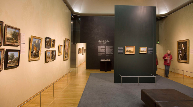 Ein großer Raum voller Gemälde an den Wänden, der zwei Personen auf dem Boden, eine Couch, einen Tisch mit verschiedenen Gegenständen, eine Tafel mit dem Text "Karel du Jardin Ausstellung in der Nationalgalerie" und eine Decke mit Lampen zeigt.