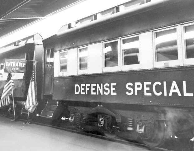 Schwarzes und weißes Foto eines Zuges mit der Aufschrift "Defense Special" auf den Eisenbahnschienen, mit zwei Fahnen links und einer Decke darüber.