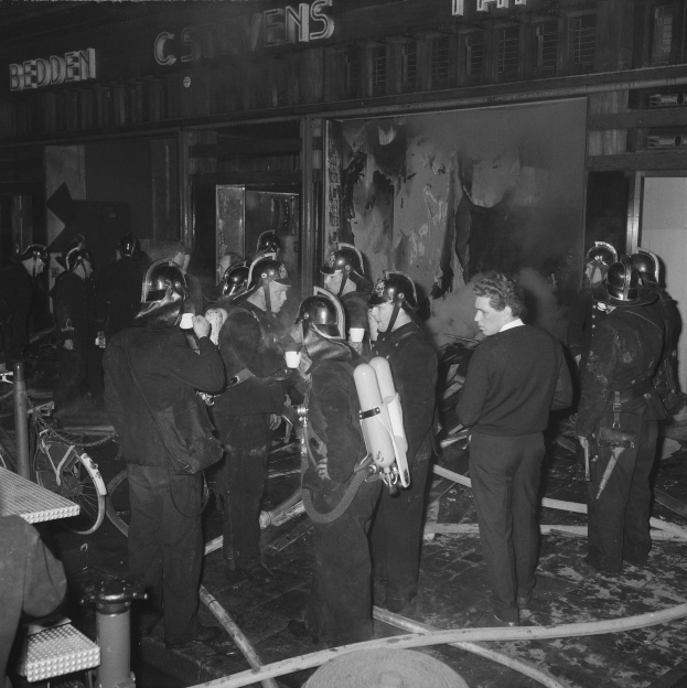 Schwarzes Foto von Feuerwehrmännern vor einem Gebäude, die Helme tragen und Luftzylinder tragen, mit einem Fahrrad und einem Tisch mit Gegenständen in der Nähe.