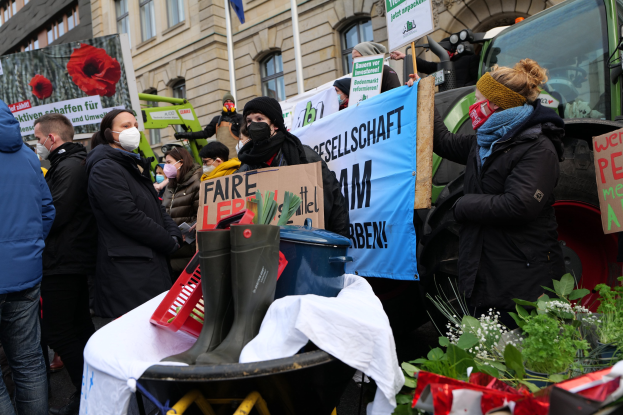 Eine Gruppe von Menschen mit Masken und Schildern protestiert vor einem Lastwagen, im Hintergrund sind ein Tisch, Pflanzen und Gebäude zu sehen.