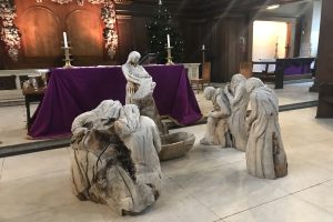 Krippe in einer Kirche mit Statuen, ein dekorierter Tisch mit Kerzen, Stühle, ein geschmückter Weihnachtsbaum und eine Hintergrundwand.
