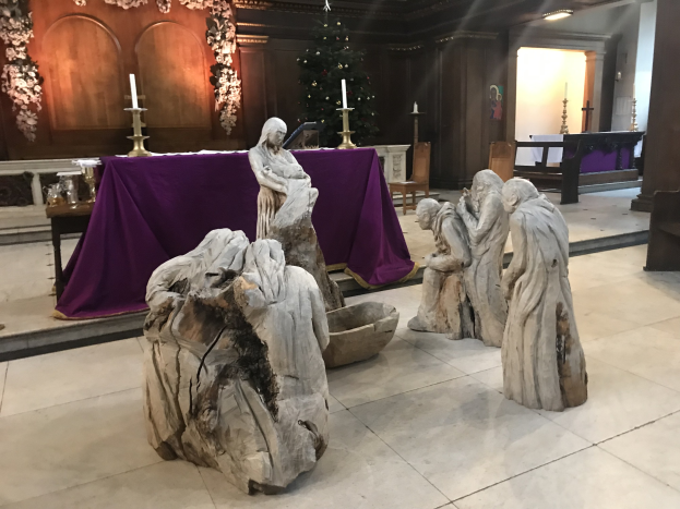 Krippe in einer Kirche mit Statuen, ein dekorierter Tisch mit Kerzen, Stühle, ein geschmückter Weihnachtsbaum und eine Hintergrundwand.
