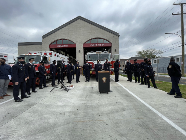 Gruppe von Menschen in Caps, die neben einem Feuerwehrauto auf einem Podium während einer Feuerwachen-Einweihungszeremonie stehen, mit Gebäuden, Bäumen, Strommasten, Laternen und Gras im Hintergrund.