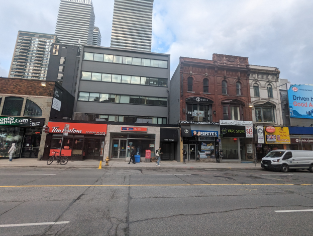 Stadtstraßenszene mit hohen Gebäuden, fahrenden Fahrzeugen, Fußgängern, einem Fahrrad, einem Hydranten und Werbetafeln für Mietwohnungen unter einem bewölkten Himmel.