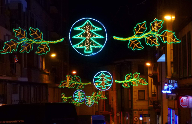 Eine städtische Straße, geschmückt mit festlichen Weihnachtslichtern, die Blätter und andere dekorative Elemente aufweist, mit Fahrzeugen auf der Straße und Gebäuden auf beiden Seiten.