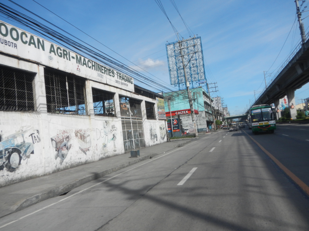 Eine Straße in einer Stadt mit Fahrzeugen, eine Brücke auf der rechten Seite, Gebäude, Strommästele mit Käblein und ein bewölkter Himmel; an einem Gebäude an der Straße befindet sich Graffiti.