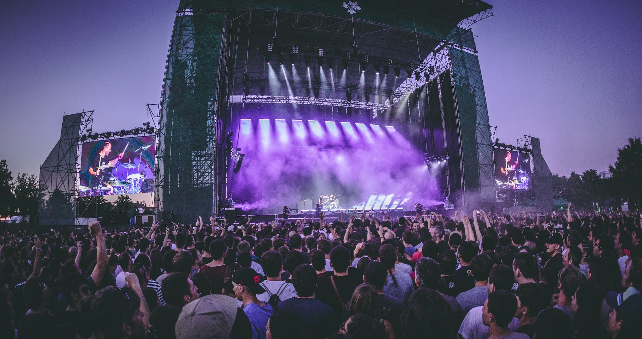 Eine große Menschenmenge auf einem Musikfestival schaut auf der Bühne mit Instrumenten und Bildschirmen, die vor einem Gebäude, Bäumen und einem klaren blauen Himmel aufgebaut sind.