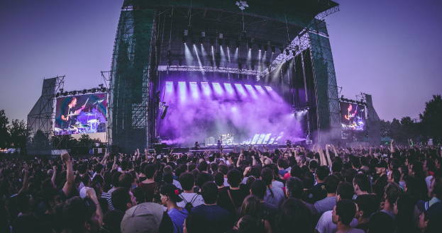 Eine große Menschenmenge auf einem Musikfestival schaut auf der Bühne mit Instrumenten und Bildschirmen, die vor einem Gebäude, Bäumen und einem klaren blauen Himmel aufgebaut sind.