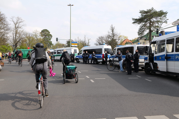 Eine Gruppe von Menschen, die auf Fahrrädern eine Straße entlangfahren, gesäumt von Polizeiwagen und anderen Fahrzeugen, einige tragen Helme und Taschen, andere schieben Kinderwagen, mit Bäumen, Laternen, Verkehrsampeln, Verkehrsschildern, Gebäuden und einem bewölkten Himmel im Hintergrund.