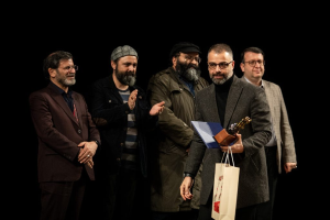 Eine Gruppe von Menschen auf einer Bühne, mit einer Frau in der Mitte, die eine Flagge hölt, einer Person mit einer Kamera links und einem Banner im Hintergrund mit der Aufschrift "Berlin as Fuck 2019".