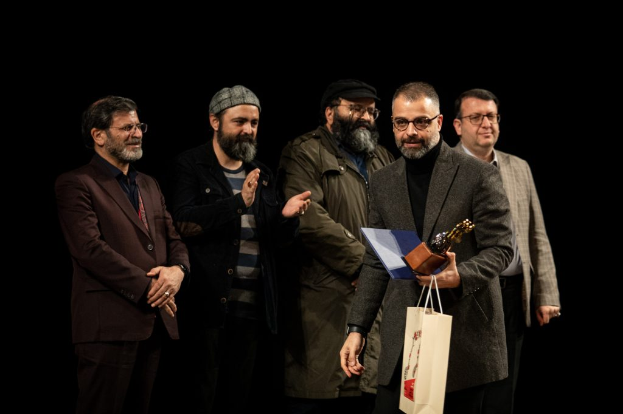 Eine Gruppe von Menschen auf einer Bühne, mit einer Frau in der Mitte, die eine Flagge hölt, einer Person mit einer Kamera links und einem Banner im Hintergrund mit der Aufschrift "Berlin as Fuck 2019".