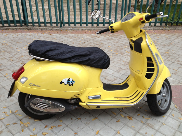 Ein gelbes Vespa GTS 300 Super ABS-Scooter, der auf einer Straße geparkt ist, mit einem schwarzen Tuch darüber, einem Geländer, Gras und Menschen im Hintergrund.