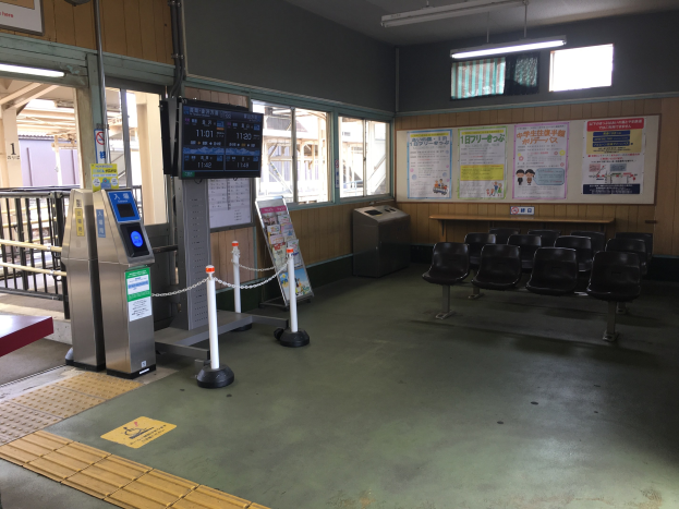 Ein belebter Bahnhofseingang mit Sitzreihen, einem Service-Schalter, Ticketautomaten, Kettenzäunen, Informationsanzeigen, Wandplakaten, Deckenbeleuchtung und Geländern.