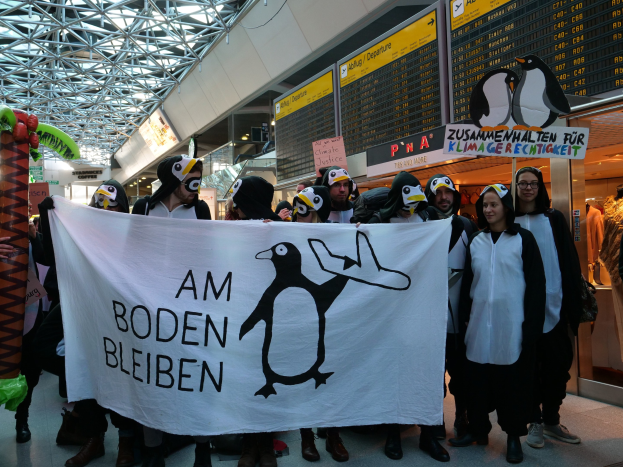 Gruppe von Menschen in Pinguinkostümen mit einem Banner vor einer Wand mit Pinguin-Plakaten, Schaufensterpuppen in Kleidern und diversen Gegenständen im Hintergrund.
