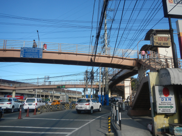 Eine Brücke über eine vielbefahrene Straße mit Fahrzeugen, Verkehrskegeln, Pfählen, Schildern, Fußgängern, Strommasten mit Drähten, Gebäuden und einem klaren blauen Himmel.