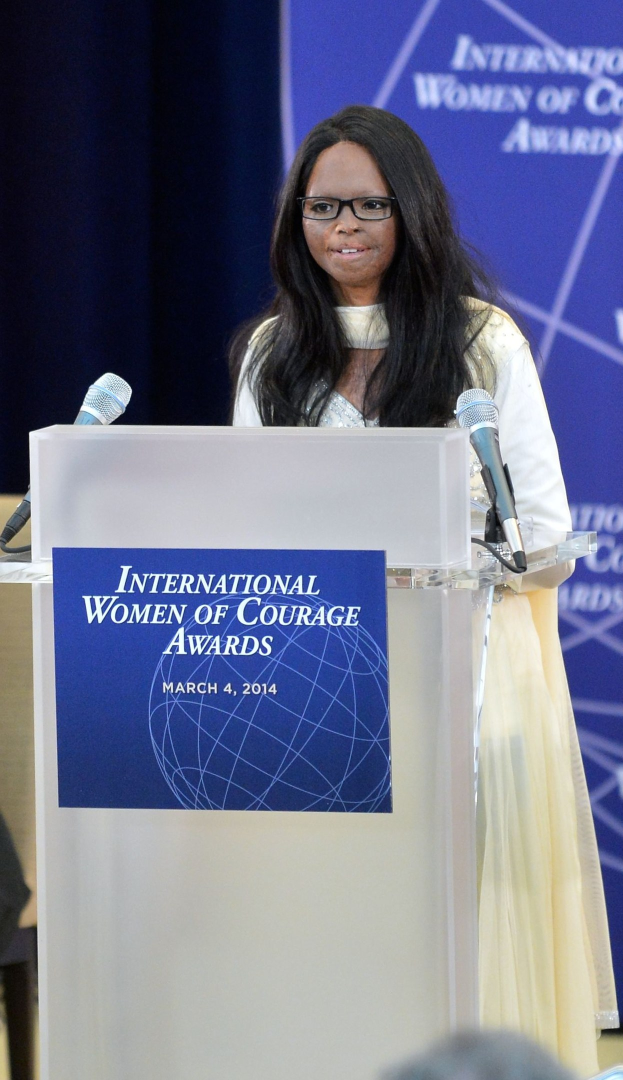 Eine Frau In Einem Weißen Kleid Und Brille Steht An Einem Podium Mit Zwei Mikrofonen Und Redet Vor Einer Menge, Mit Einer Blauen Flagge Im Hintergrund, Die 'Internationale Frauen Des Mutes Awards 2014' Bezeichnet.