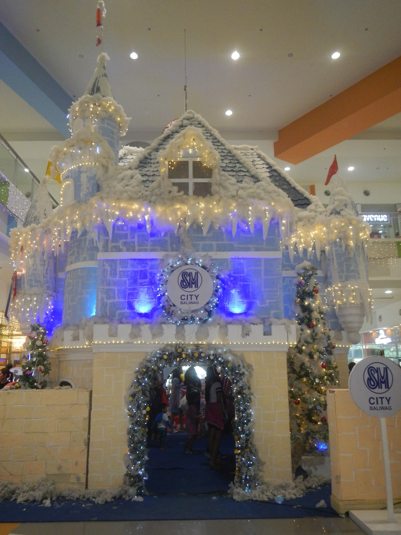 Ein festliches Weihnachtsdorf in einem Einkaufszentrum mit einer Burgen-ähnlichen Struktur mit Lichtern und Dekorationen, umgeben von Menschen, mit einer Texttafel auf der rechten Seite und beleuchteten Deckenlichtern, die eine gemütliche Atmosphäre schaffen.