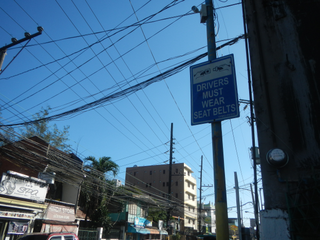 Eine Stadtstraße mit Autos, Strommasten mit Drähten, Gebäuden, Bäumen und Namensschildern, mit einem Schild an einem Strommasten, auf dem steht "Fahrer müssen den Sicherheitsgurt anlegen".