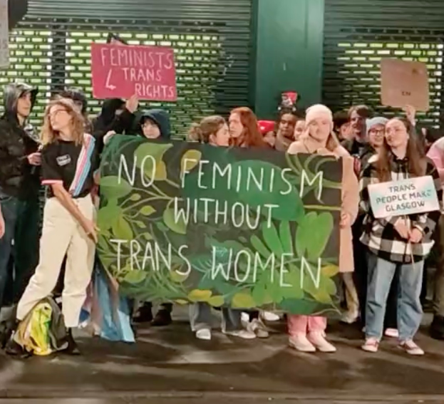 Eine Gruppe von Menschen hält ein Transparent mit der Aufschrift "Kein Feminismus ohne Trans-Frauen" und verschiedene Schilder, die in einem öffentlichen Raum mit einer Wand und einem Rollladen im Hintergrund stehen.