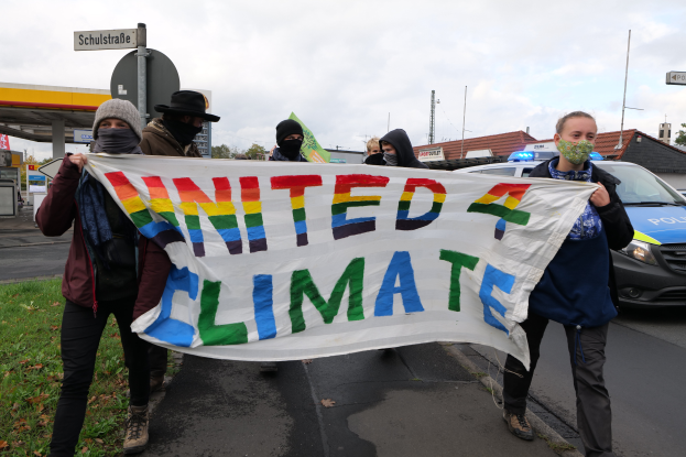 Eine Gruppe maskierter Personen mit einem "United Climate"-Schild vor einer Tankstelle, mit Fahrzeugen, Gras, Schildern, Gebäuden, Bäumen und einem bewölktem Himmel im Hintergrund.