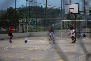 Junge Jungs spielen Basketball auf einem Outdoor-Court mit einem Zaun, Bäumen, Gebäuden und einem klaren blauen Himmel im Hintergrund.