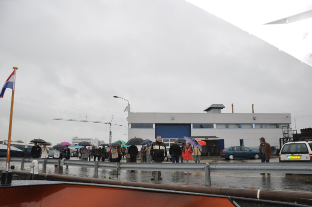 Eine Gruppe von Menschen, die draußen stehen, einige halten Schirme, mit einer Fahne an einem Mast, zwei Autos, einem Gebäude mit Fenstern, einem Turmkran, einer Straßenlaterne und einer Barrikade im Hintergrund.
